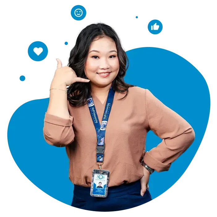 Smiling person making a call gesture, wearing a lanyard on a blue background.